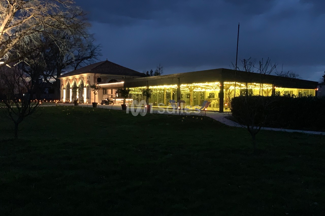 Bâtiment éclairé avec des guirlandes lumineuses, entouré d'arbres et d'une pelouse, sous un ciel sombre.