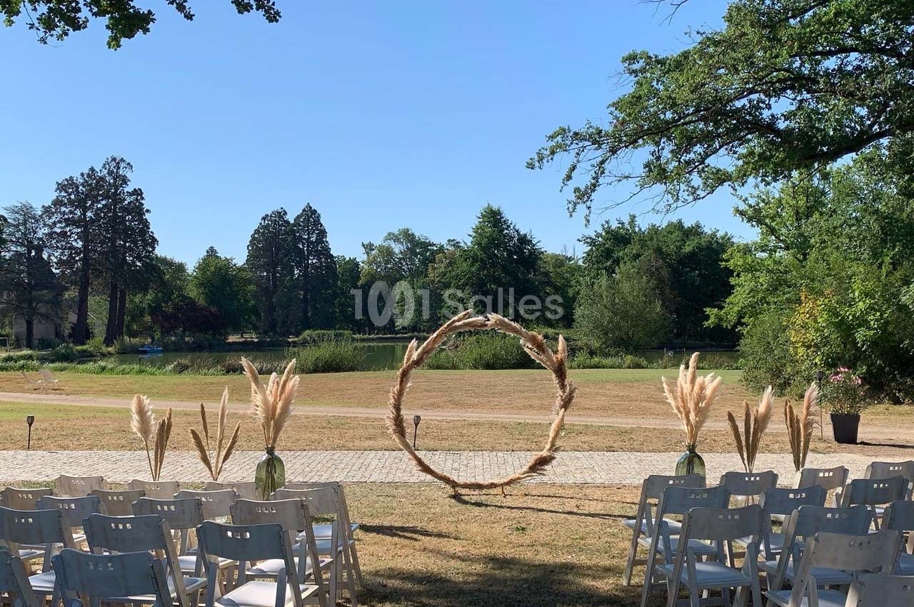 Chaises blanches alignées en extérieur face à une arche circulaire décorée, avec un lac et des arbres en arrière-plan.