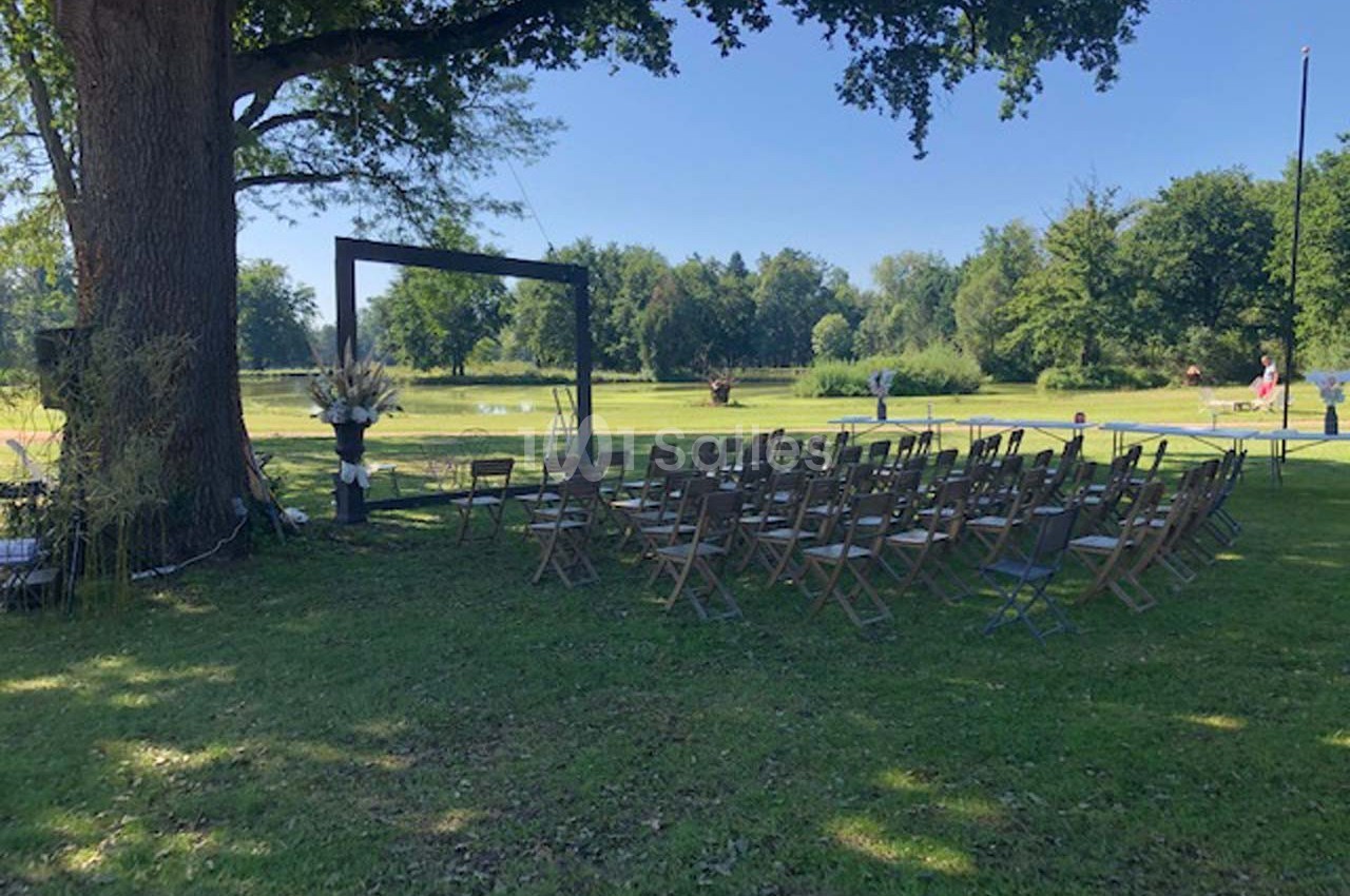 Chaises en bois disposées en rangées sous un grand arbre, formant un espace extérieur pour une cérémonie.
