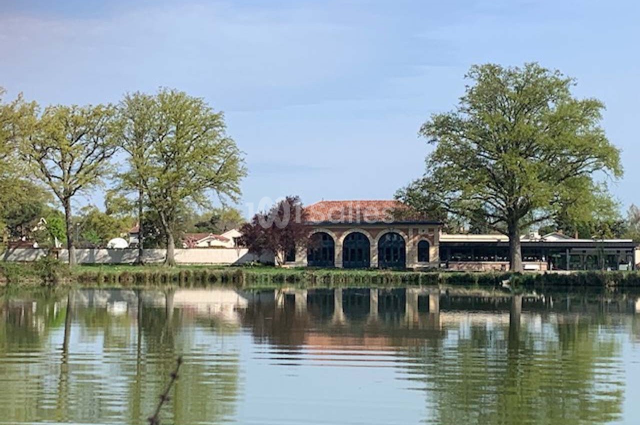 Bâtiment en pierre avec arches, situé au bord d'un étang entouré d'arbres et de végétation.