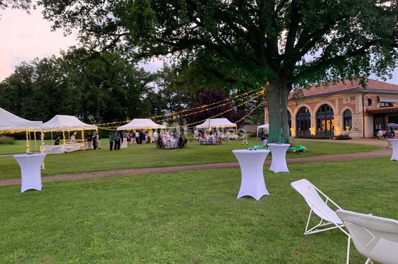Jardin avec tentes blanches, guirlandes lumineuses et tables hautes lors d'un événement en plein air au crépuscule.