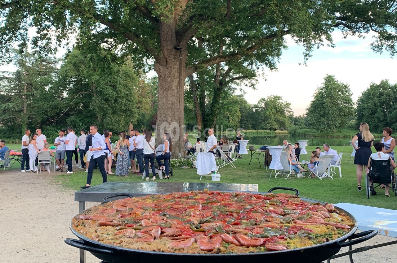 Grande poêle de paella devant un groupe de personnes rassemblées dans un jardin avec des tables et des arbres en arrière…