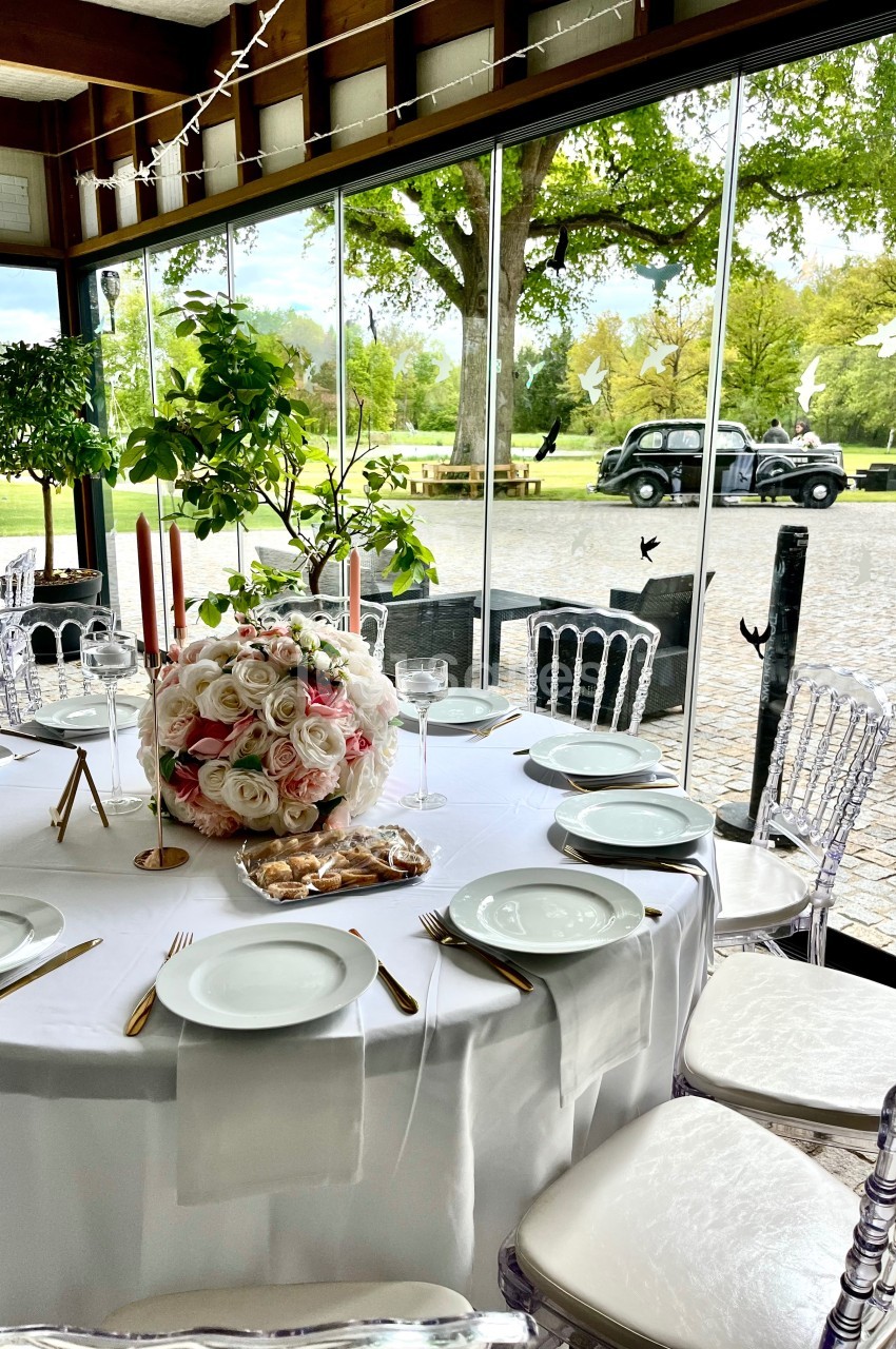 Table ronde élégamment dressée avec des assiettes blanches, des couverts dorés et un bouquet de roses, près d'une baie…