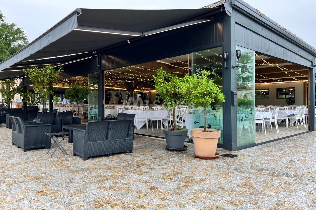 Terrasse couverte avec mobilier de jardin, entourée de plantes en pot, donnant sur une salle aménagée pour un événement.