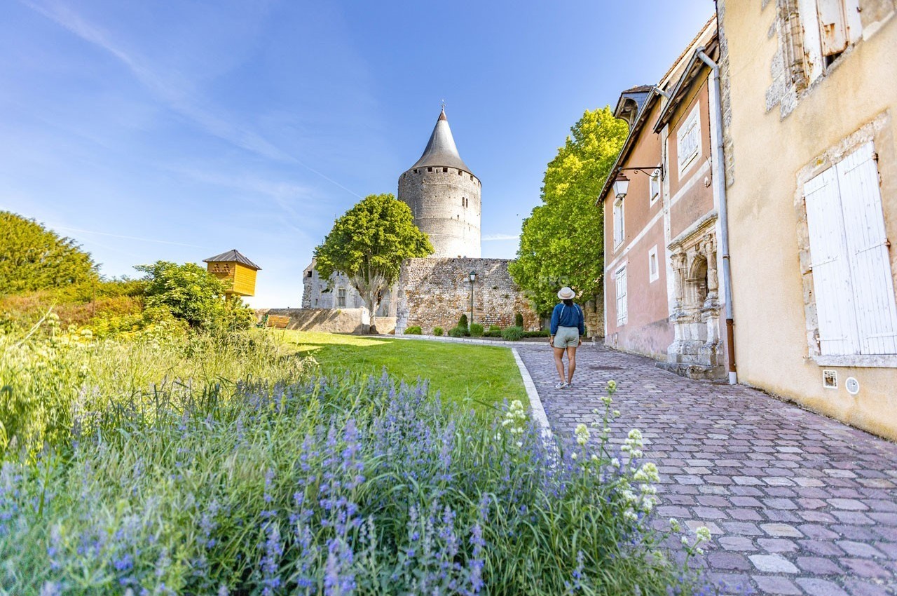 Un homme marche sur une allée pavée bordée de fleurs, menant à une tour médiévale entourée de murs et de maisons anciennes.