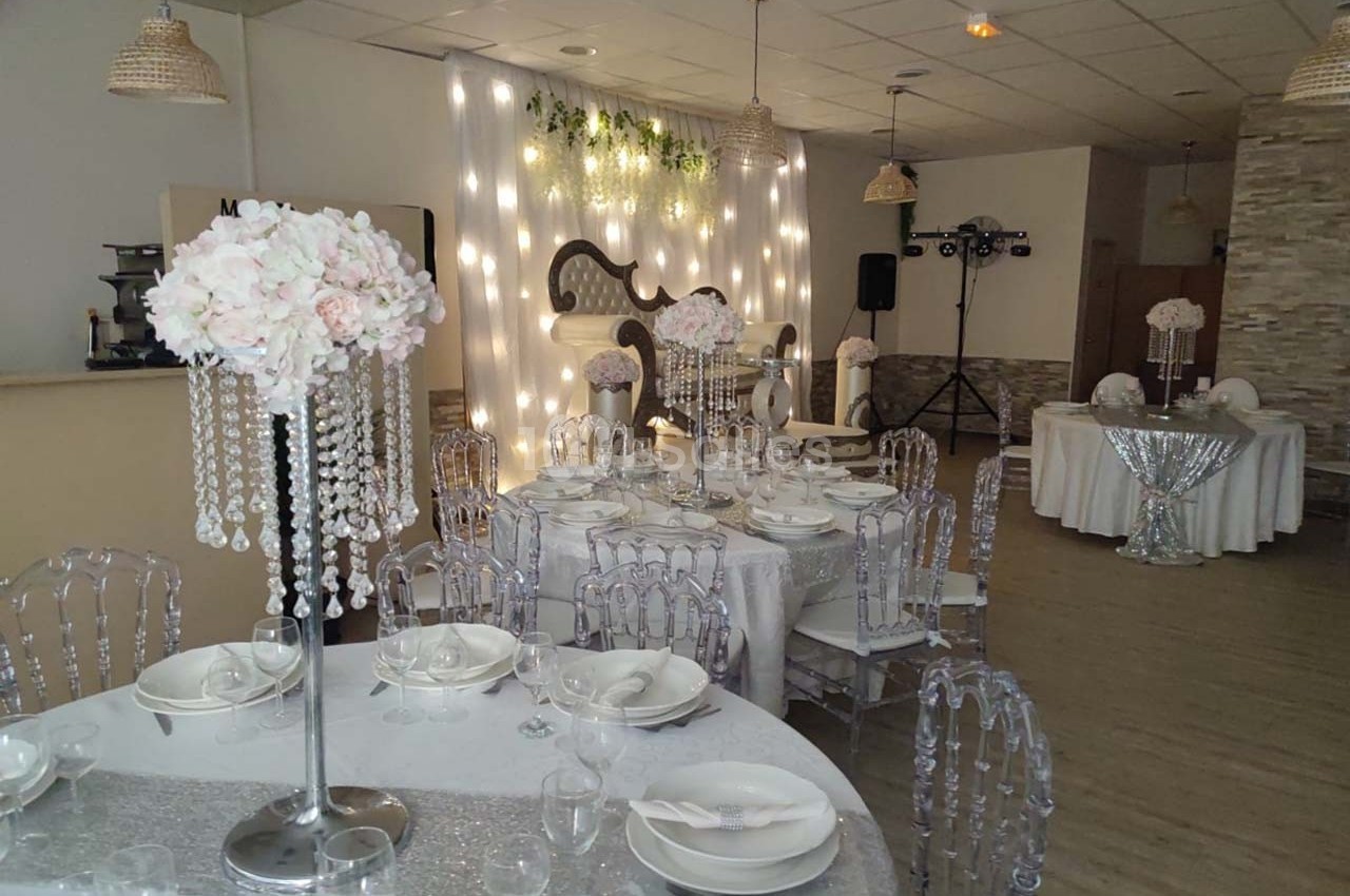 Salle de réception décorée avec des tables dressées, des chaises transparentes et des centres de table fleuris avec perles.