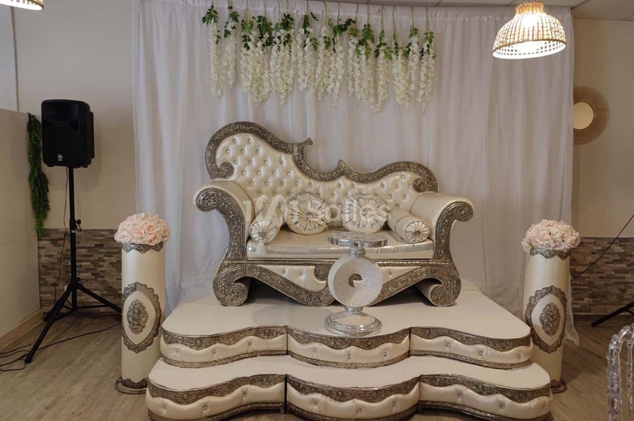 Canapé décoratif argenté et blanc sur une estrade, entouré de fleurs et d'une draperie blanche avec guirlandes suspendues.