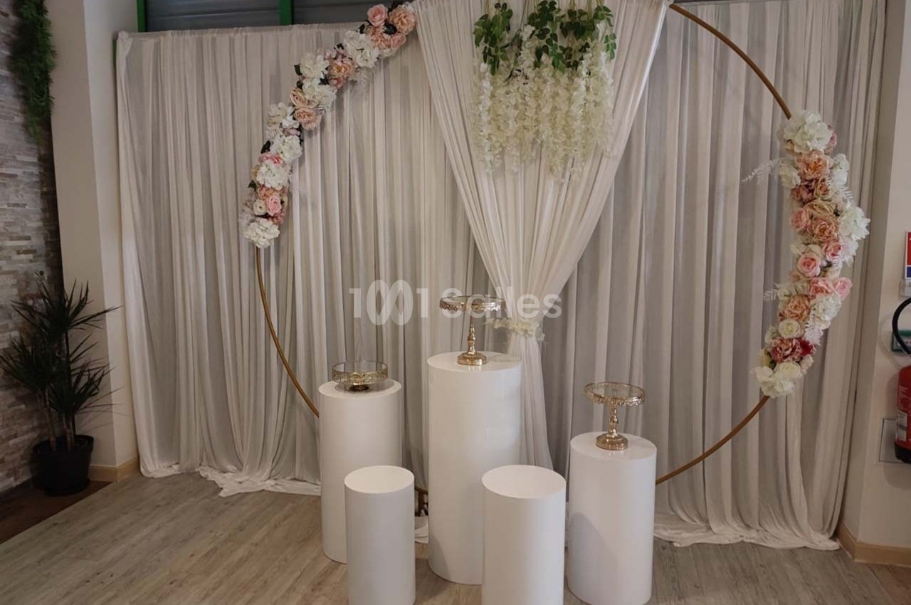Cercle doré décoré de fleurs, devant un rideau blanc plissé, avec des colonnes blanches et supports transparents.