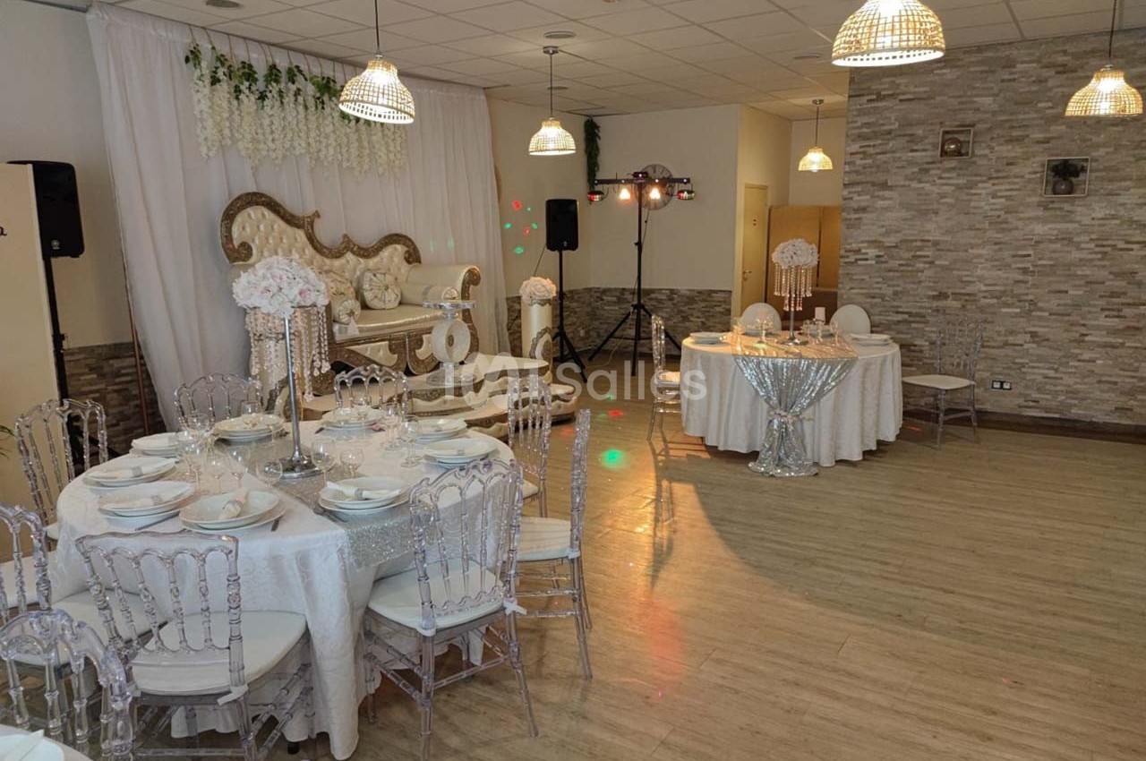 Salle de réception décorée avec des tables dressées, des chaises transparentes et un canapé orné sous un rideau blanc.