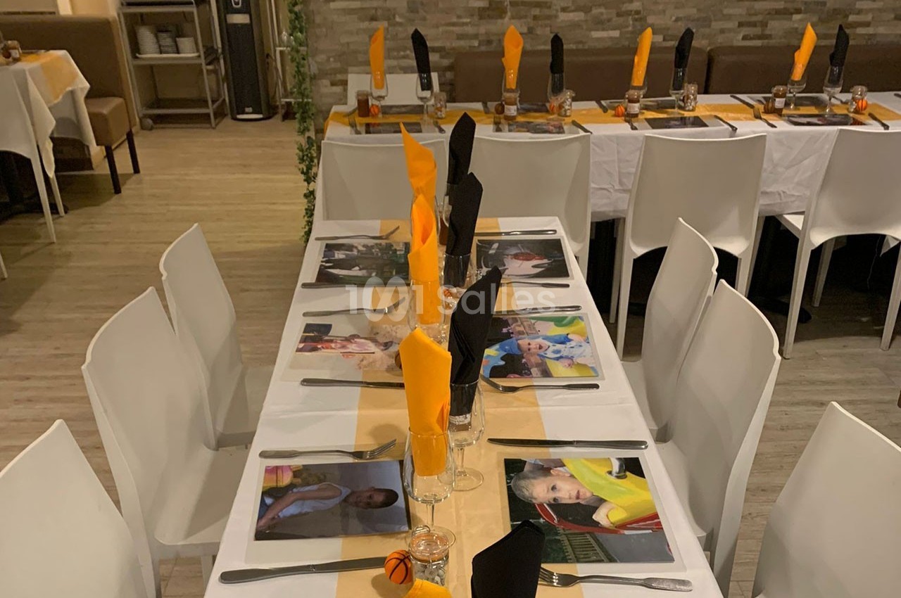 Tables dressées avec nappes blanches, serviettes orange et noires pliées, et sets de table illustrés dans une salle décorée.