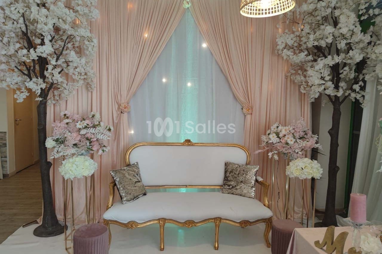 Canapé doré et blanc entouré de rideaux roses, arbres fleuris et décorations florales dans un cadre élégant.