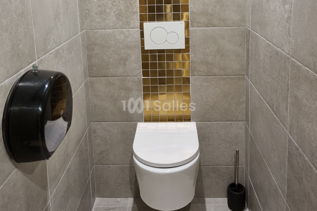 Toilettes modernes avec cuvette suspendue, carrelage gris, mosaïque dorée et accessoires noirs.