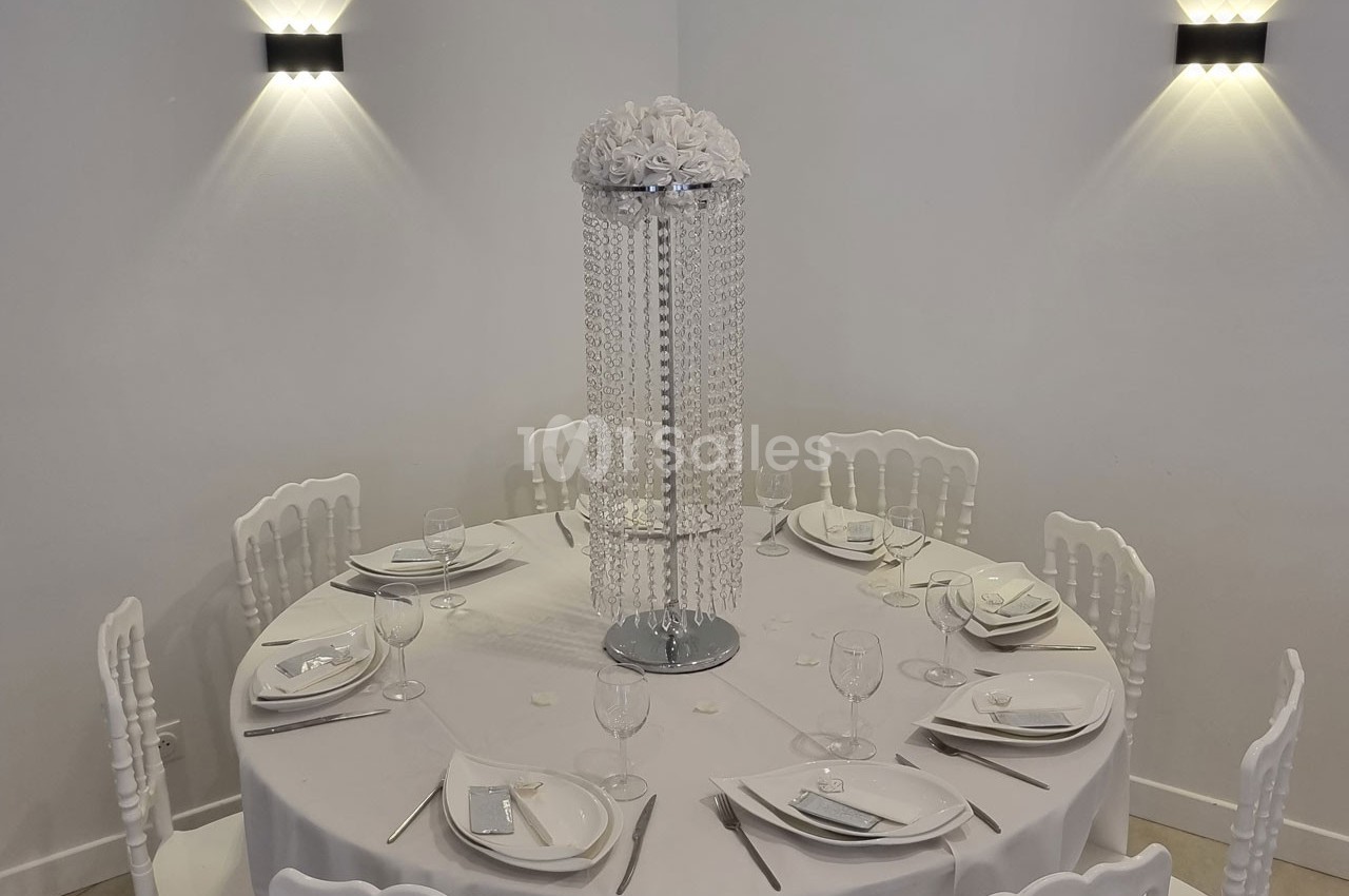 Table ronde décorée pour un événement, avec nappes blanches, vaisselle élégante et centre de table orné de perles et fleurs.