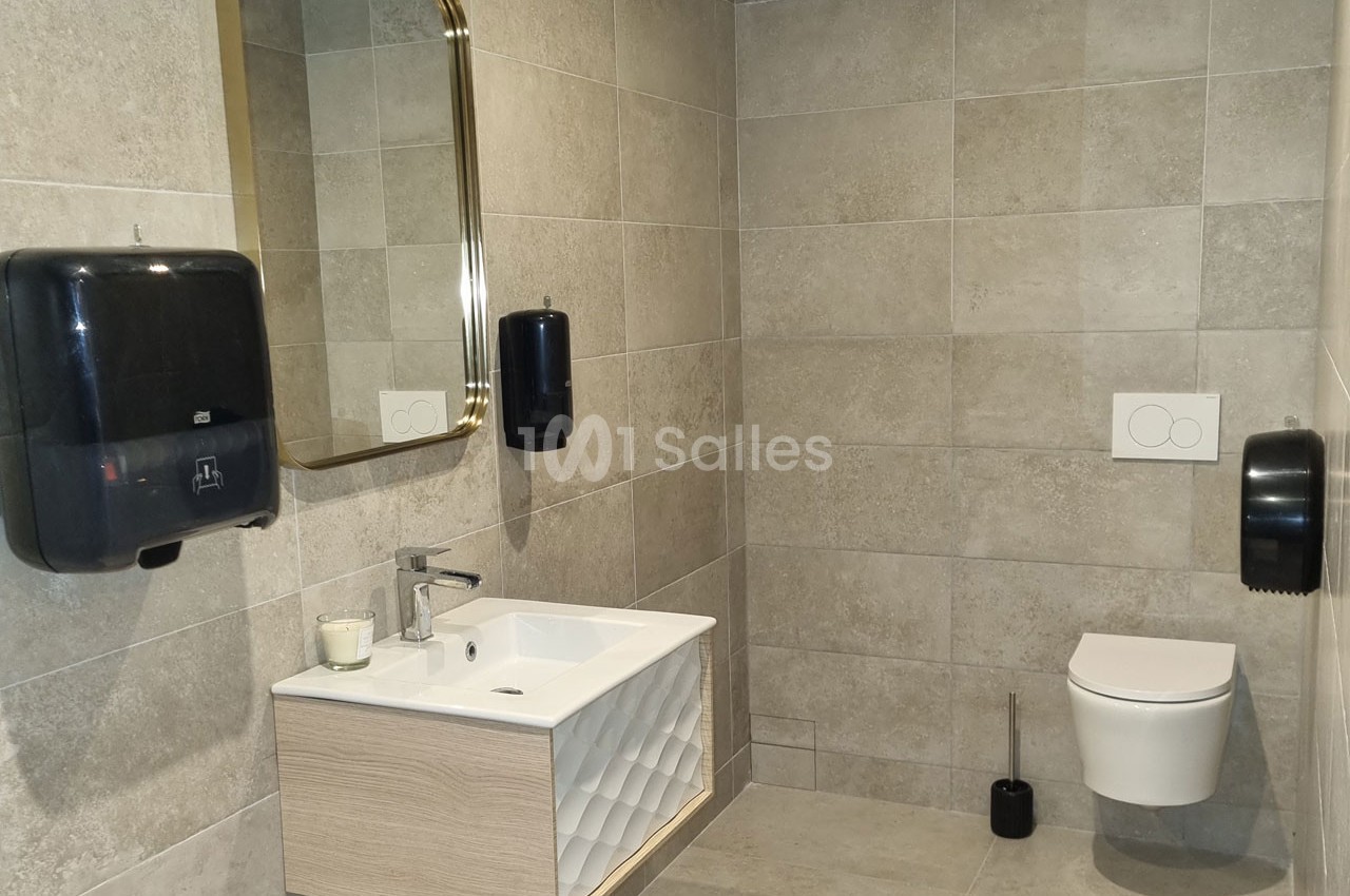 Salle de bain moderne avec lavabo, miroir doré, distributeurs de savon, toilettes suspendues et carrelage beige.