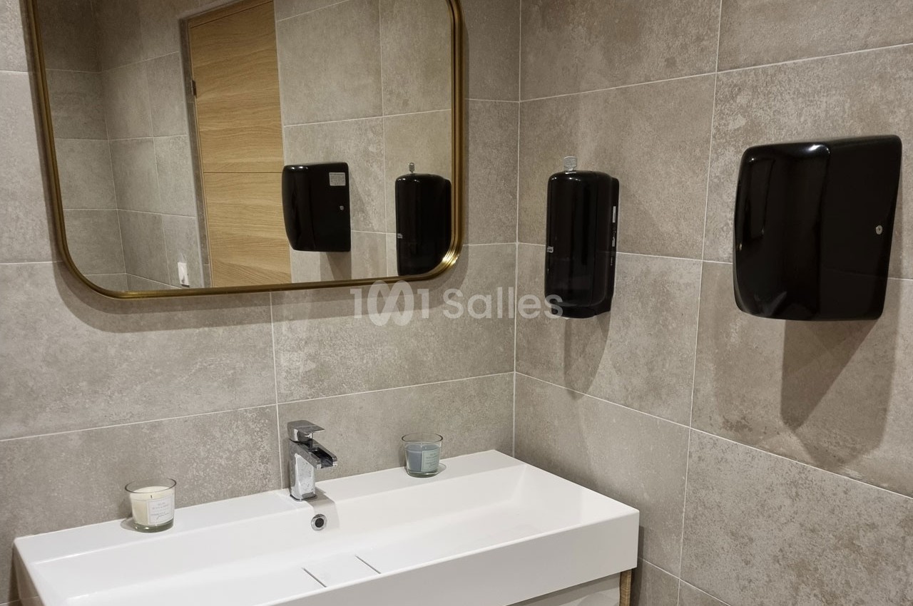 Lavabo blanc avec miroir rectangulaire doré, distributeurs noirs et murs en carrelage beige.