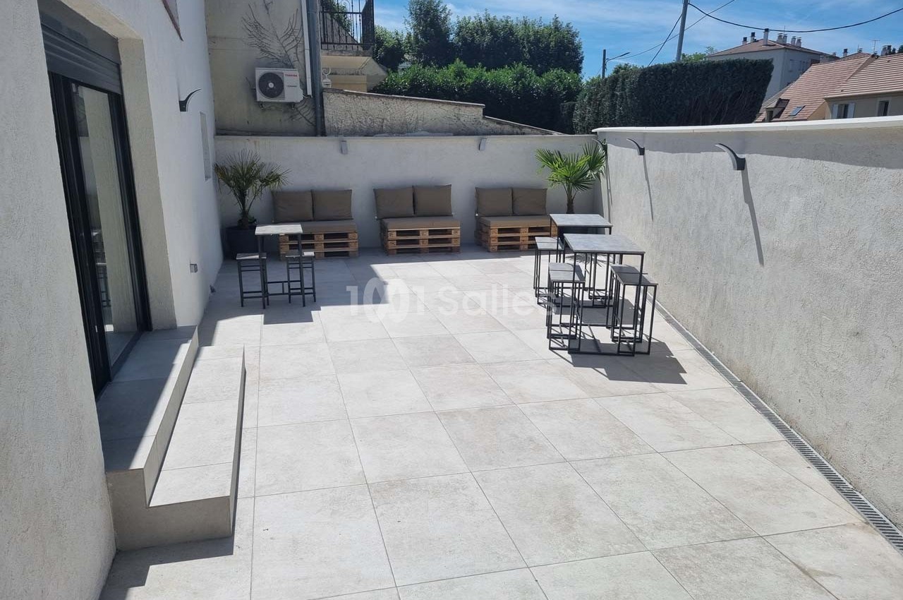 Terrasse ensoleillée avec sol carrelé, mobilier en palettes et tables en métal, entourée de murs clairs.