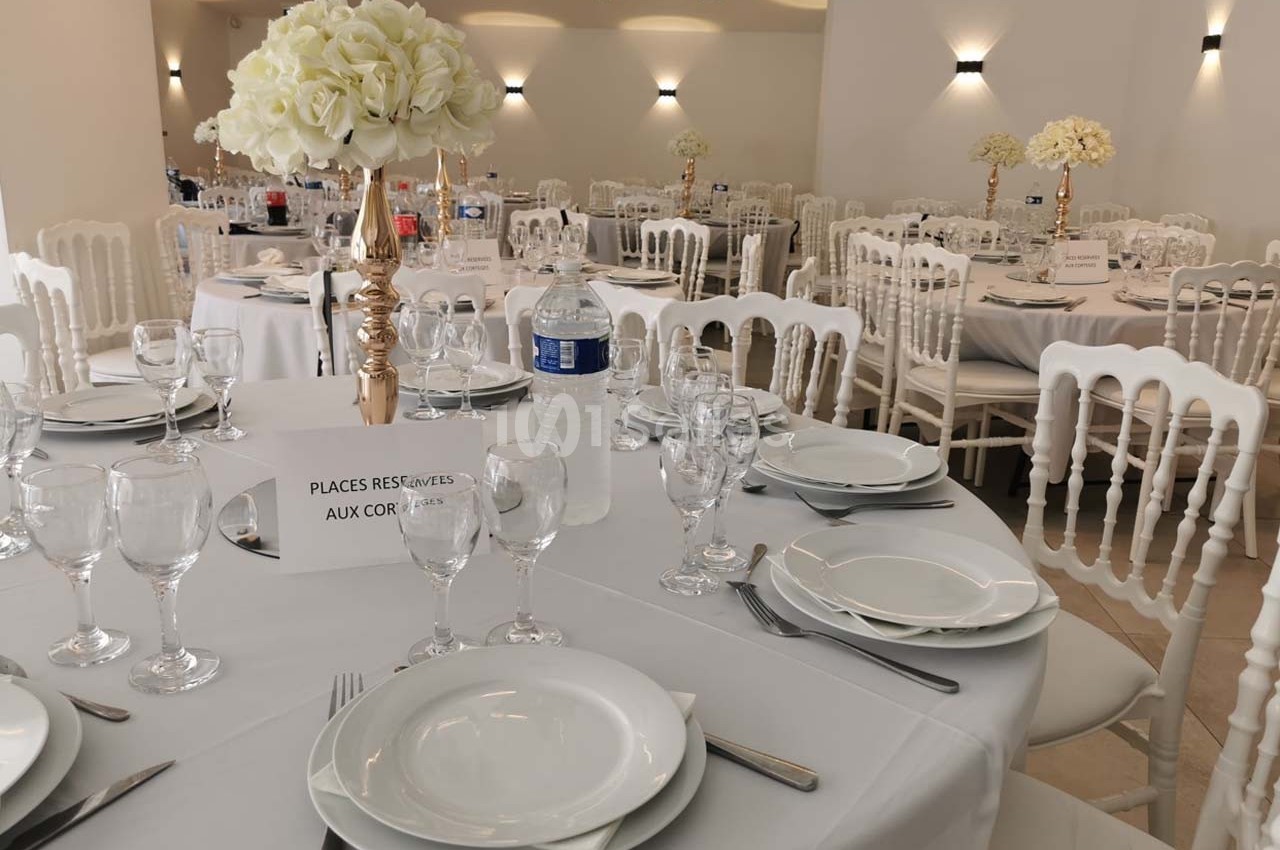 Salle de réception décorée avec des tables rondes dressées, ornées de bouquets de fleurs blanches.
