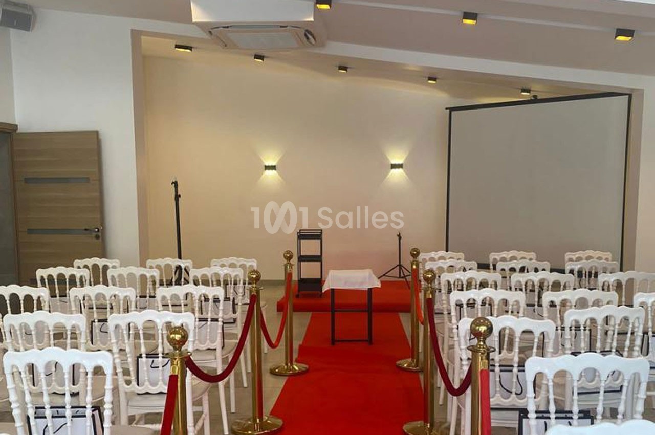 Salle aménagée avec des chaises blanches, tapis rouge, pupitre et écran de projection, prête pour un événement.