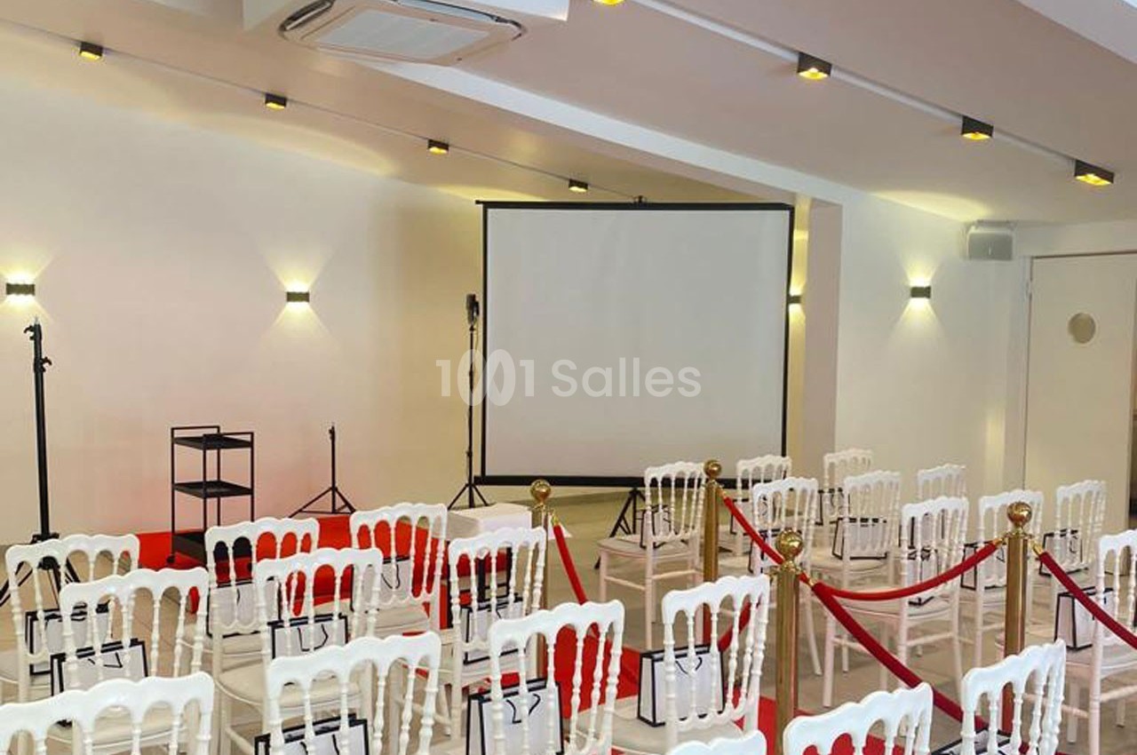 Salle aménagée avec des chaises blanches, un écran de projection, des micros et des cordons rouges délimitant l'espace.