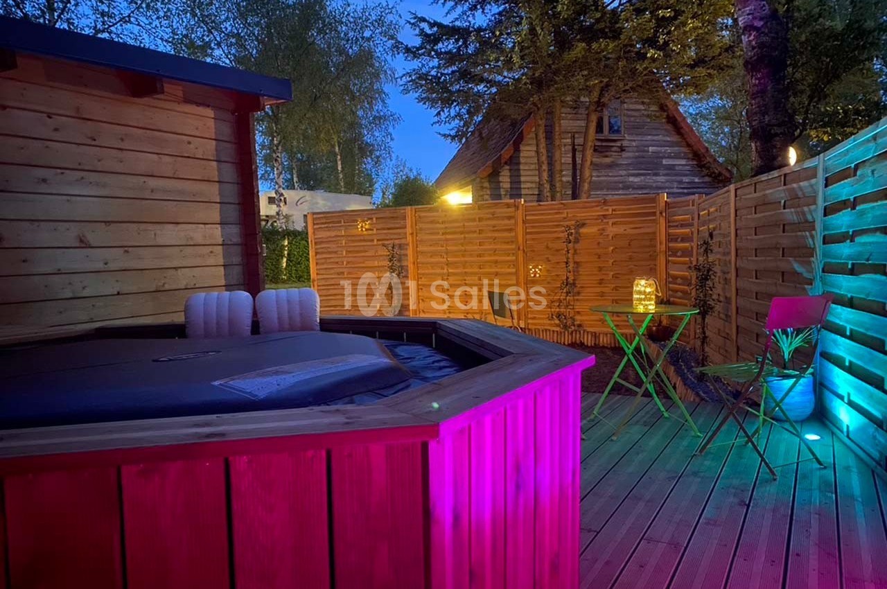 Terrasse en bois avec jacuzzi éclairé, table et chaises colorées, entourée de clôtures et végétation en soirée.
