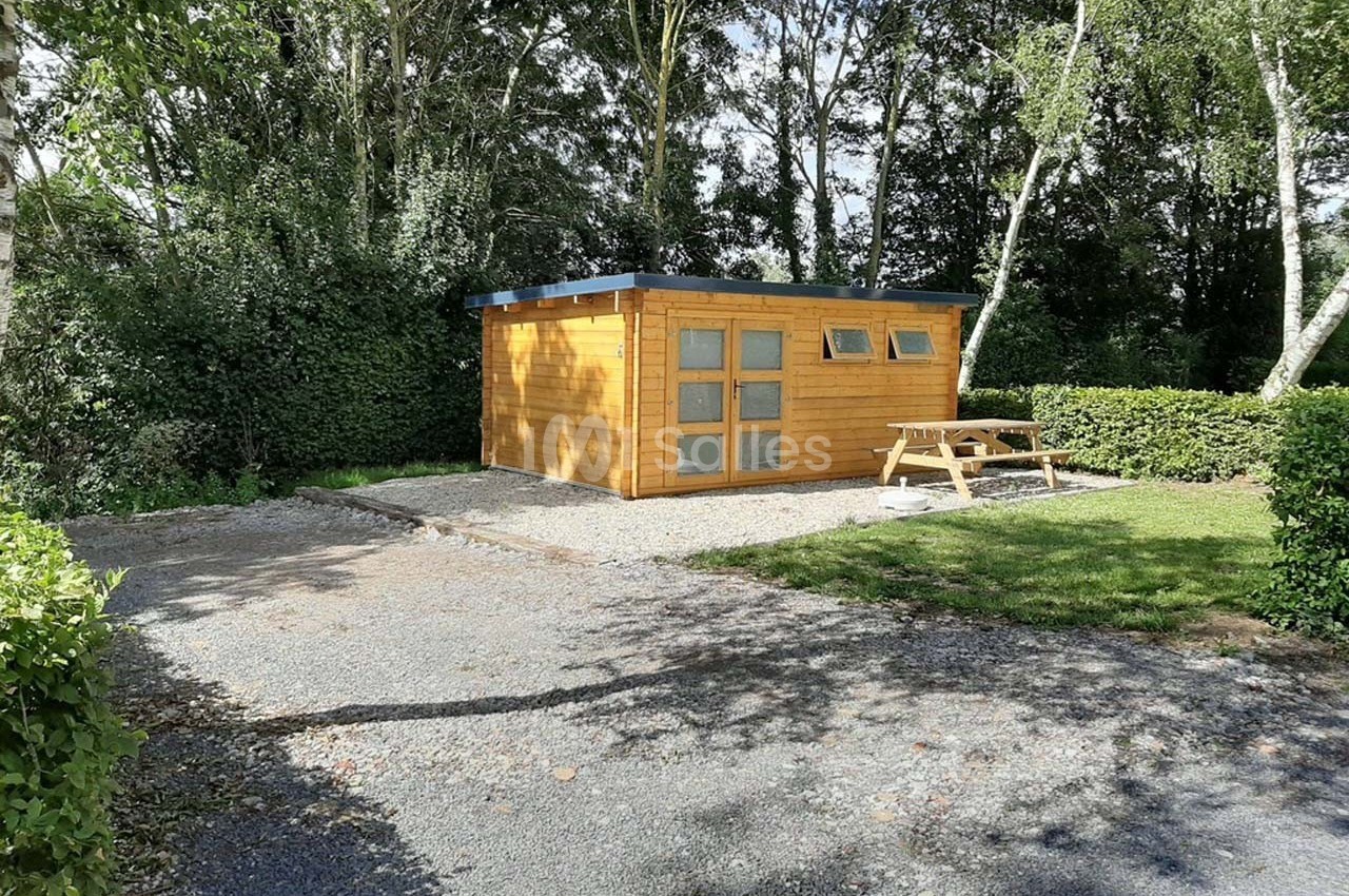 Abri en bois avec fenêtres, entouré de végétation, situé sur un terrain gravillonné avec une table de pique-nique.