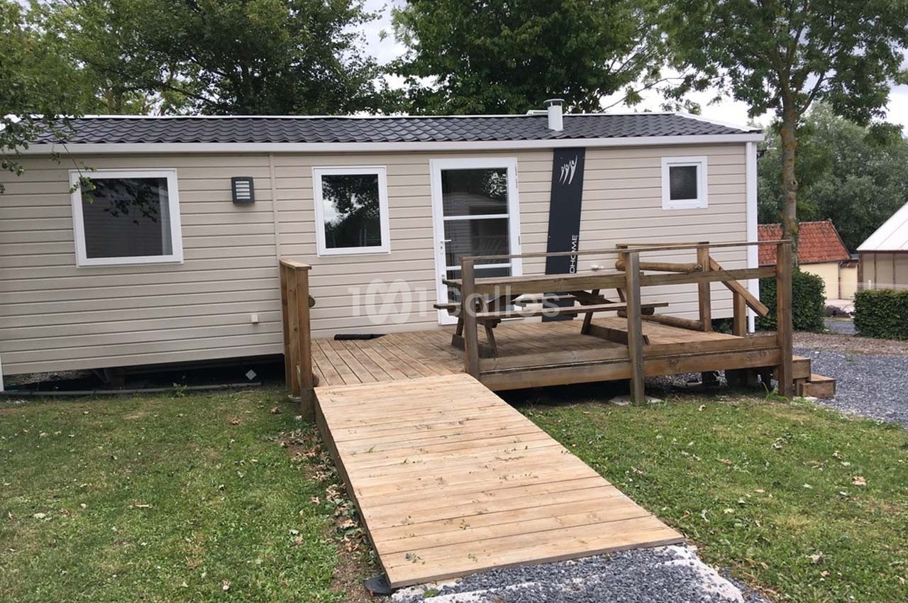 Mobil-home beige avec terrasse en bois accessible par une rampe, entouré de pelouse et d'arbres.