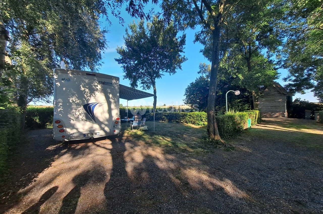 Camping-car stationné sur un emplacement ombragé avec table et chaises, entouré d'arbres et de haies.