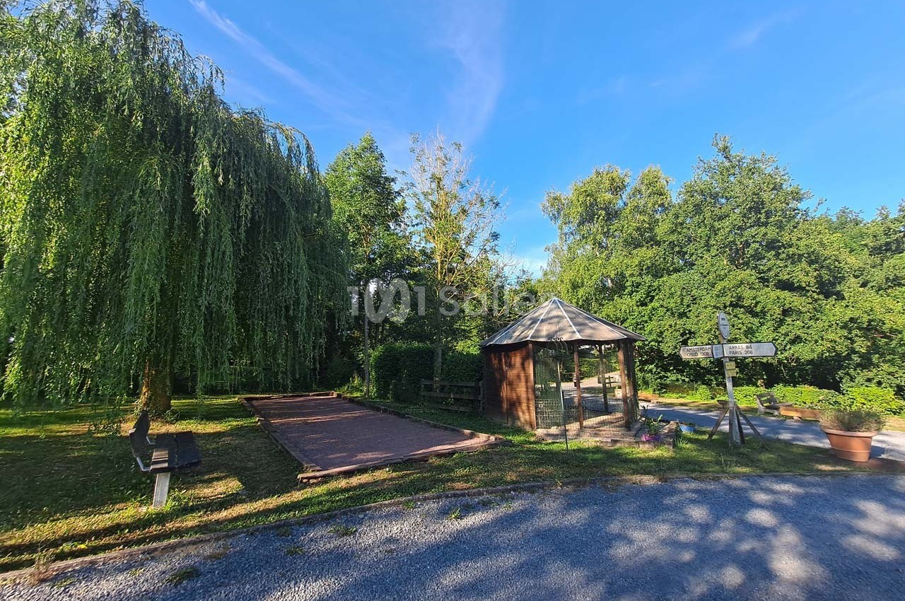 Kiosque en bois près d'un terrain de pétanque, entouré d'arbres et d'un chemin ensoleillé.