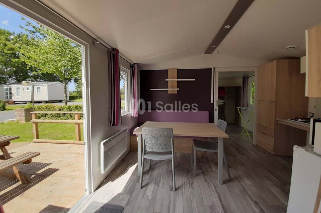 Intérieur lumineux d’un mobil-home avec table, chaises, cuisine équipée et vue sur une terrasse en bois.