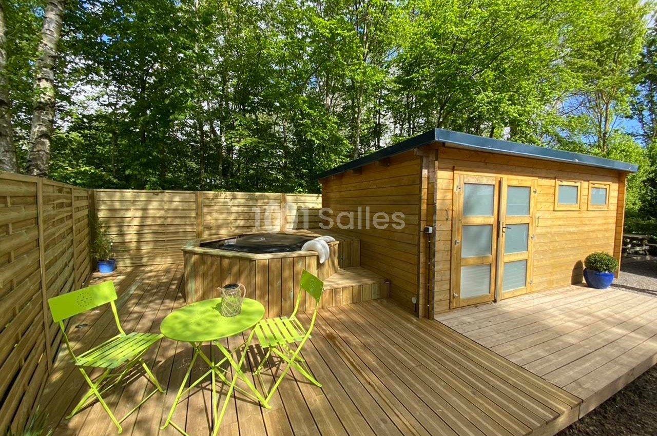 Terrasse en bois avec un jacuzzi, une table et des chaises vertes, entourée de verdure et d'une cabane en bois.