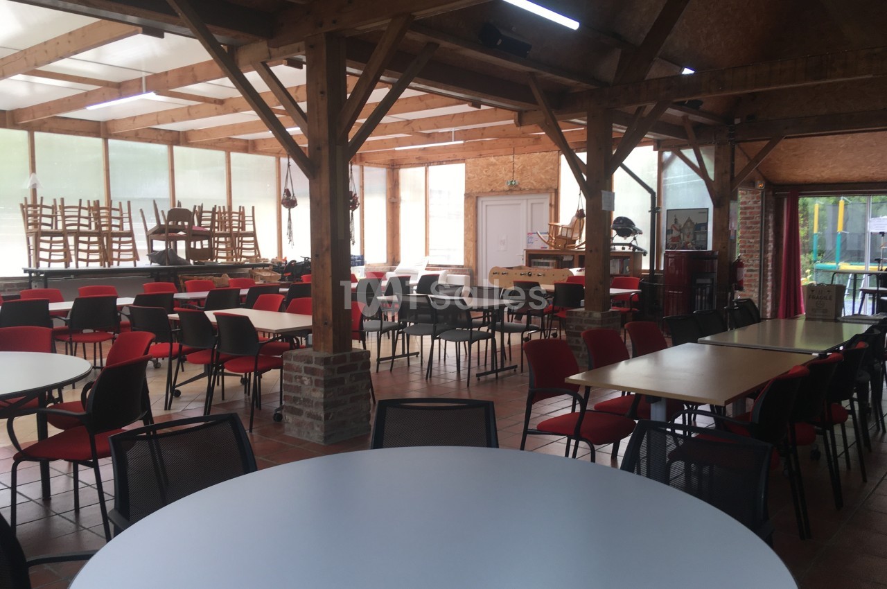 Salle lumineuse avec des tables rondes et rectangulaires, chaises empilées et structure en bois apparent.