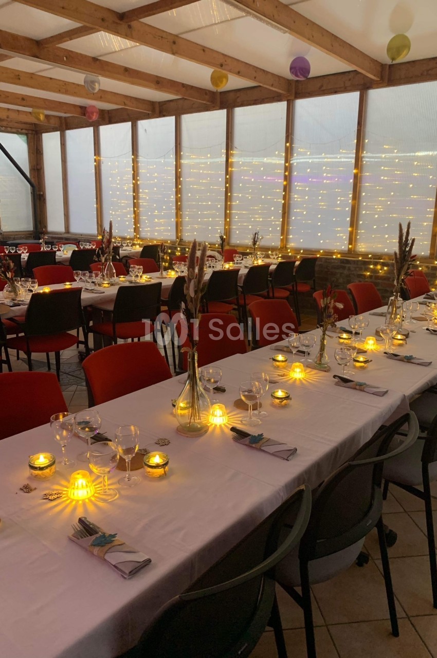 Salle décorée pour un repas, avec longues tables dressées, bougies allumées et guirlandes lumineuses en arrière-plan.