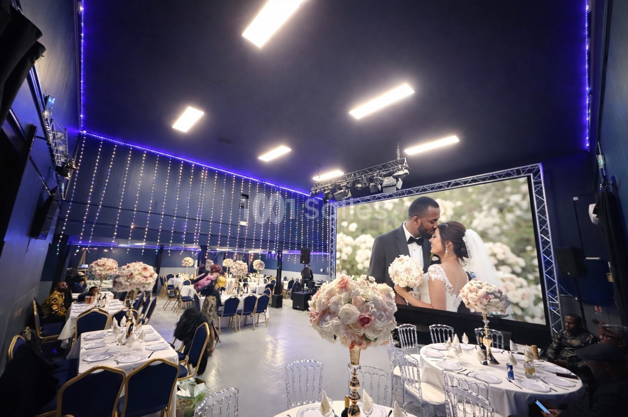 Salle de réception décorée avec des tables dressées, éclairages suspendus et un écran affichant un couple de mariés.