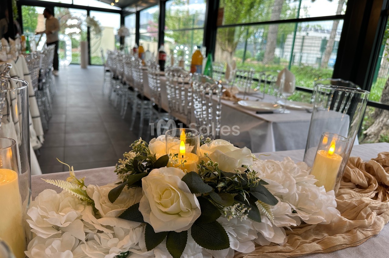 Centre de table floral avec roses blanches et bougies LED, dans une salle de réception lumineuse avec tables dressées.