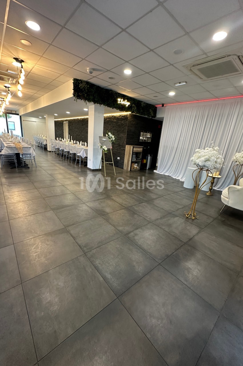Salle de réception moderne avec tables dressées, sol en carrelage gris, éclairage suspendu et décor floral.