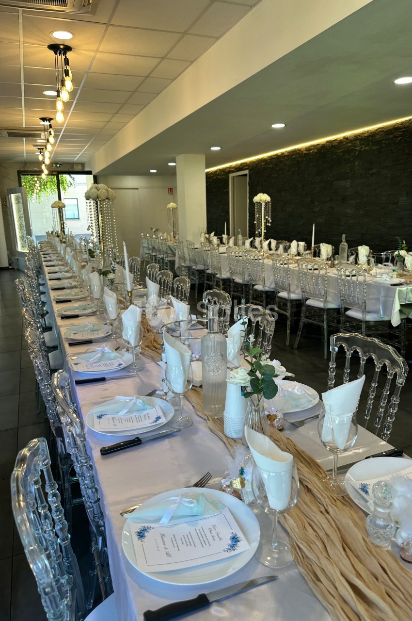 Salle de réception décorée avec de longues tables alignées, nappes blanches, chaises transparentes et centres de table…