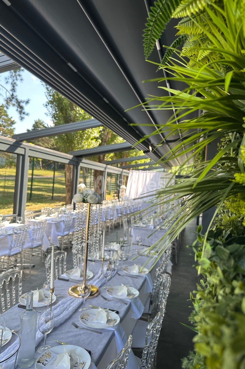 Salle de réception lumineuse avec tables dressées, chaises transparentes et décorations végétales, vue sur un espace…