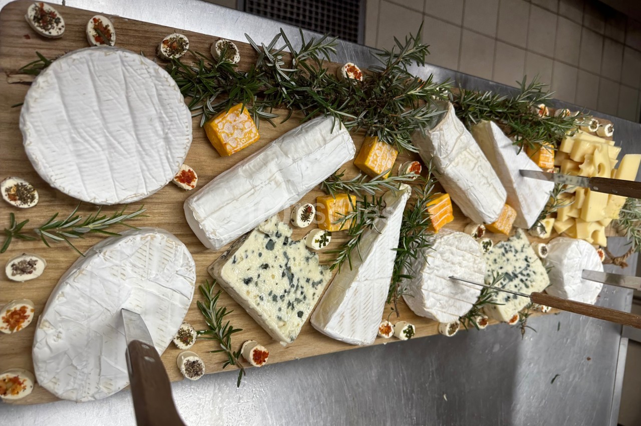 Plateau en bois garni de fromages variés, herbes fraîches et petites garnitures, présenté sur une table en cuisine.