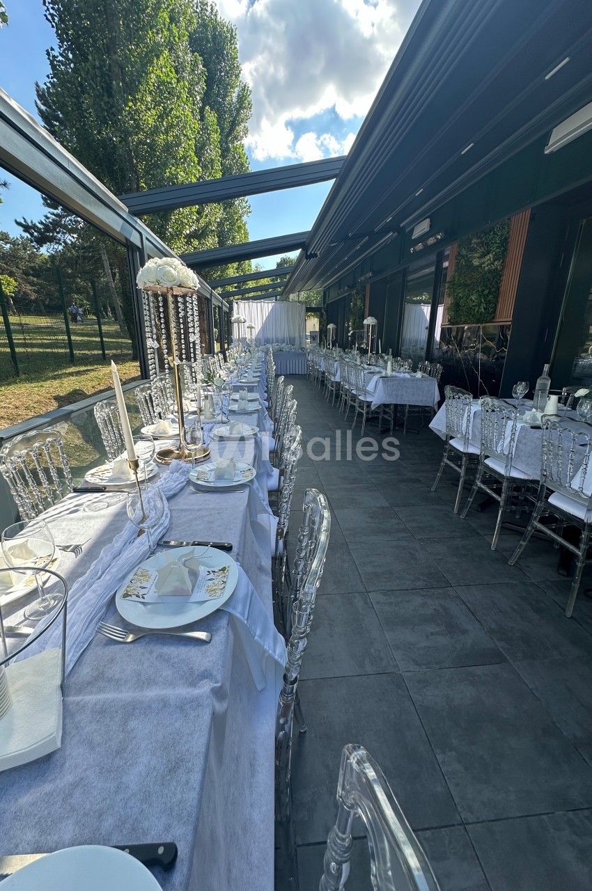 Salle de réception extérieure avec tables dressées, nappes blanches et chaises transparentes sous une verrière.