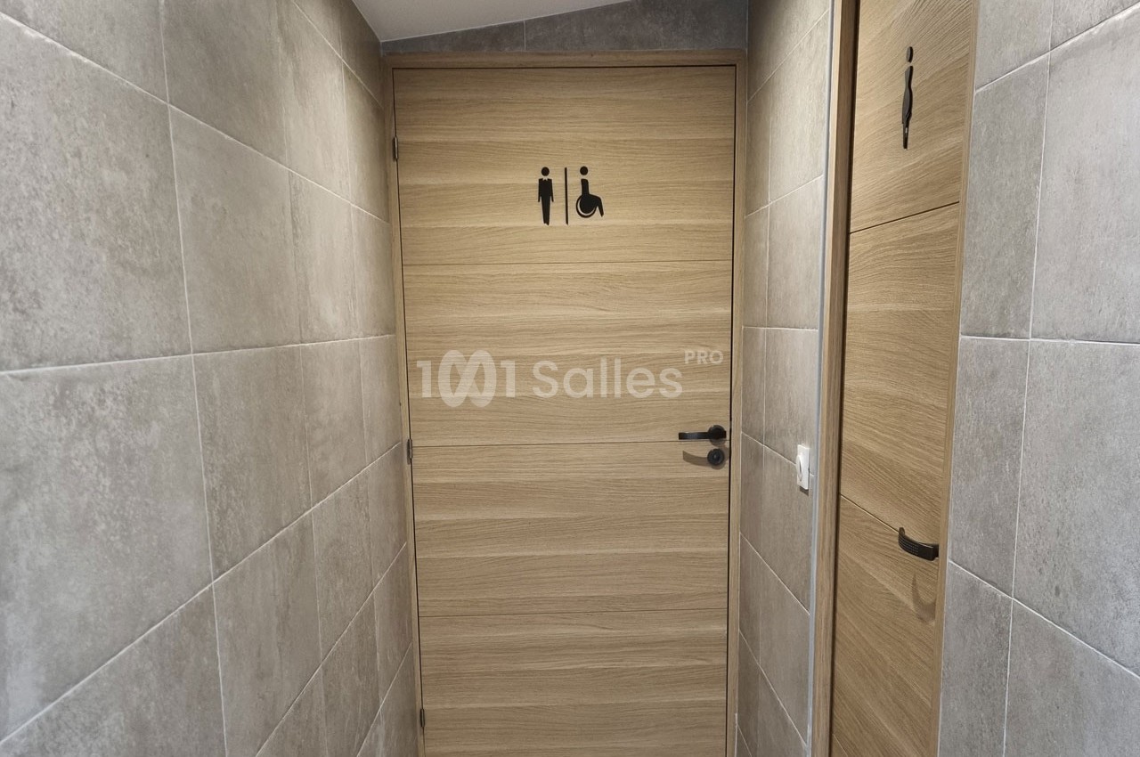 Porte de toilettes accessibles avec pictogrammes homme, femme et fauteuil roulant, entourée de murs carrelés gris.