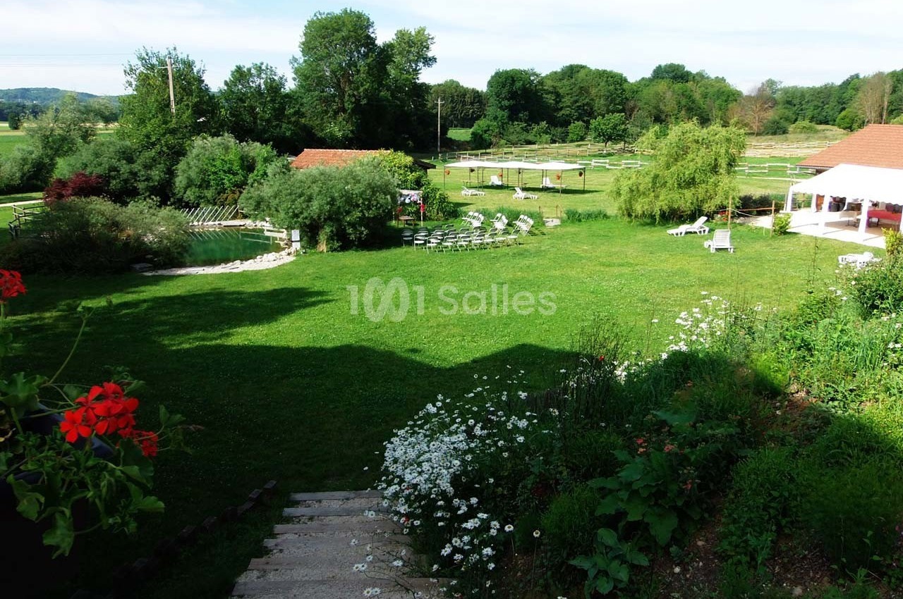 Jardin verdoyant avec pelouse, fleurs, chaises disposées en cercle et bâtiments en arrière-plan par temps ensoleillé.