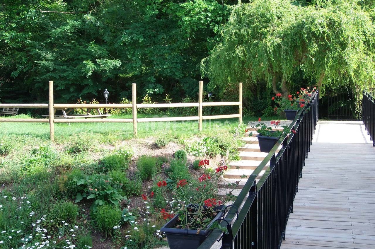 Passerelle en bois bordée de jardinières fleuries, menant à un espace vert avec arbres et clôture en bois.