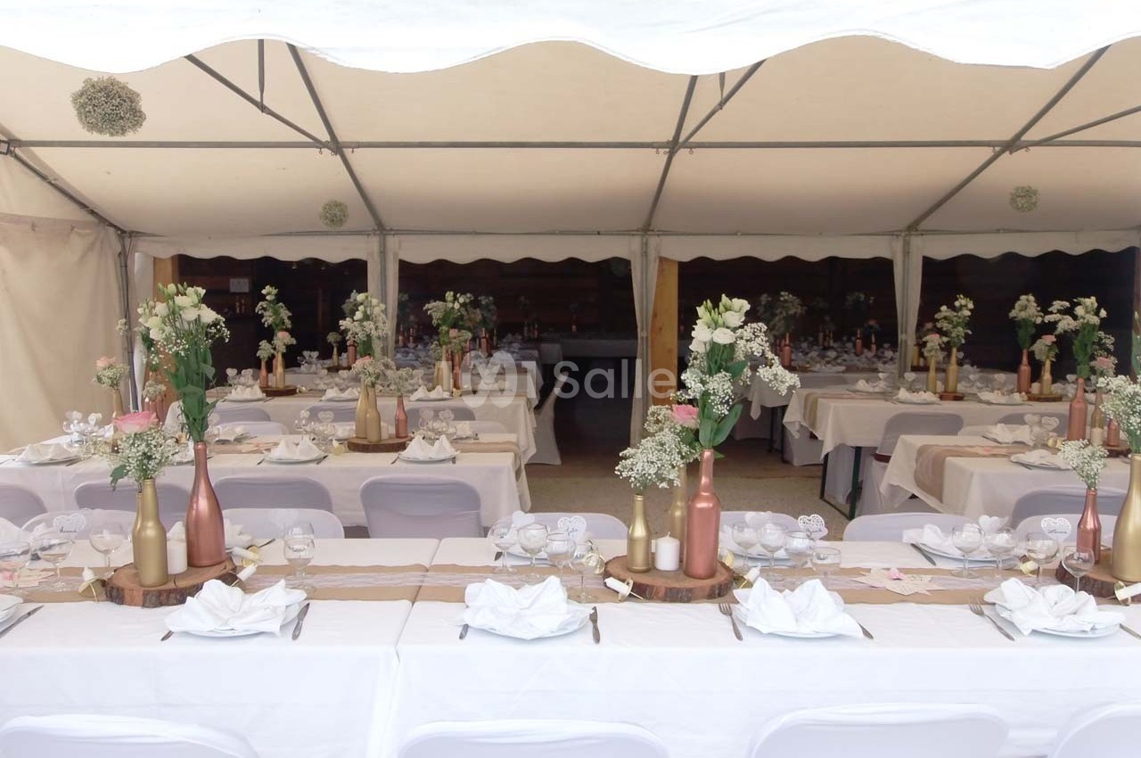 Tables décorées pour un événement sous une tente, avec nappes blanches, centres de table fleuris et vaisselle disposée.