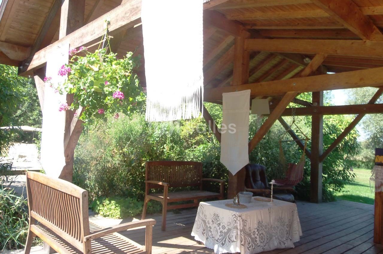 Terrasse en bois couverte avec bancs, table décorée d'une nappe blanche et plantes suspendues en fleurs.