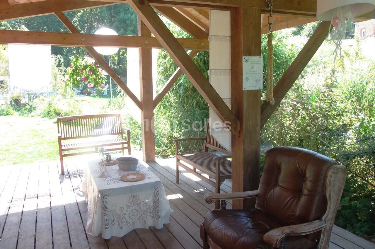 Terrasse en bois couverte avec fauteuil, bancs, table décorée et vue sur un jardin verdoyant en arrière-plan.