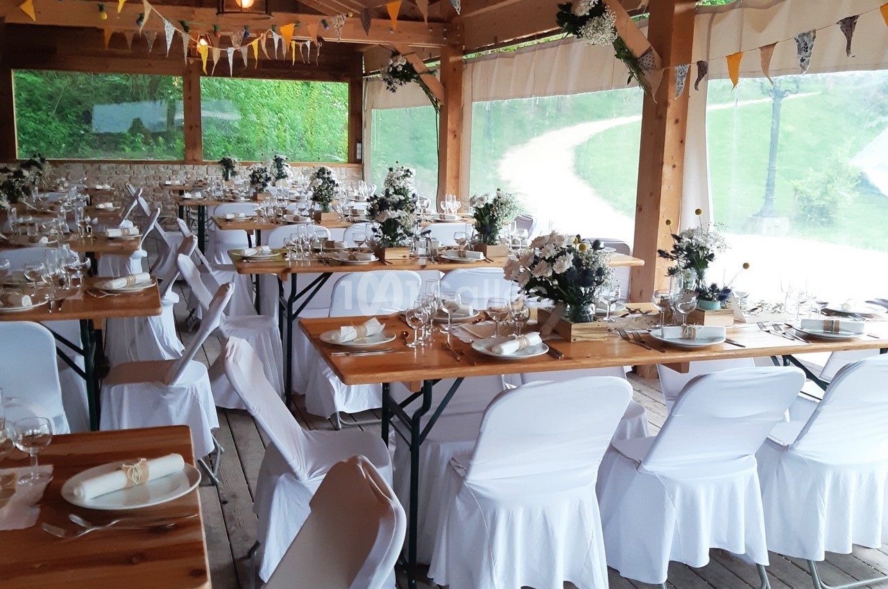 Salle de réception décorée avec des tables en bois, nappes blanches et arrangements floraux, prête pour un événement.