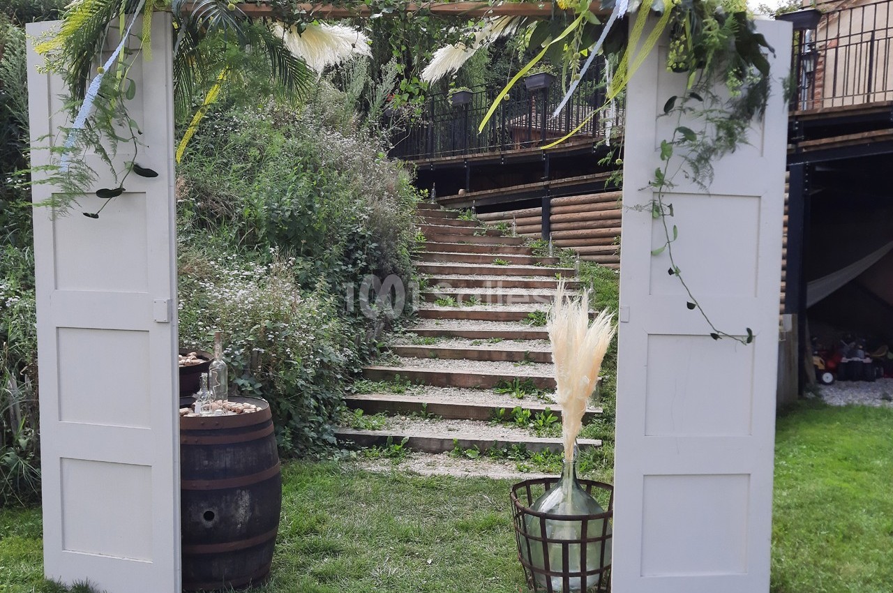 Portique décoratif en bois blanc avec feuillages et fleurs, placé devant un escalier extérieur en pierre.