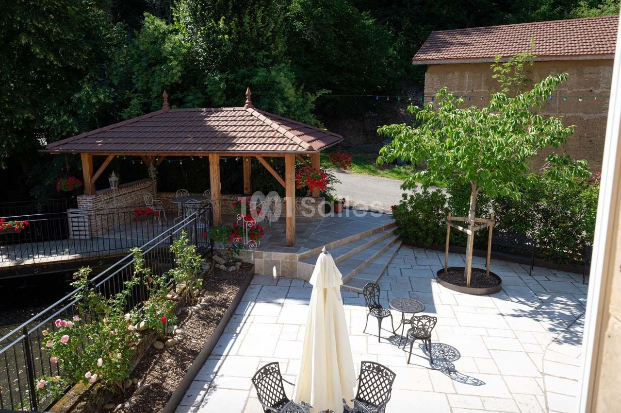 Terrasse pavée avec mobilier de jardin, pergola en bois, parterre fleuri et arbres, entourée de verdure.