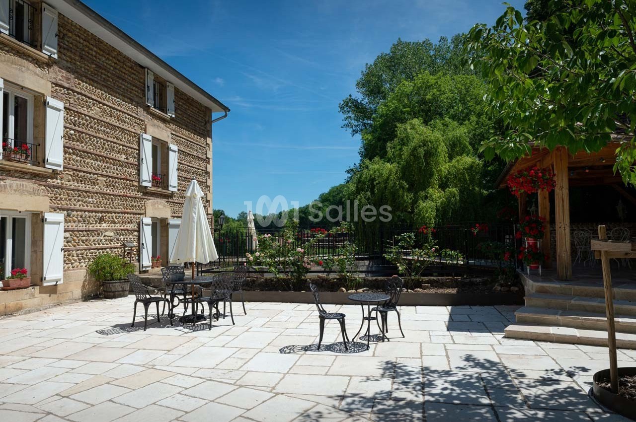 Terrasse pavée avec tables et chaises en fer forgé, entourée de verdure et d'un bâtiment en pierre avec volets blancs.