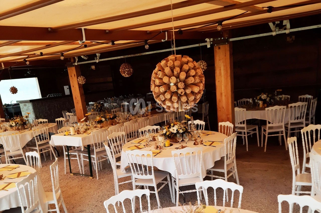 Salle décorée pour un événement, avec des tables rondes dressées, chaises blanches et suspensions en bois.